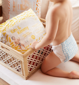 A toddler in a diaper reaching into a wooden basket containing packaged diapers.