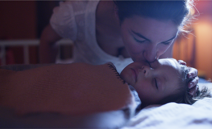 A woman kisses a sleeping child on the forehead under a blanket.