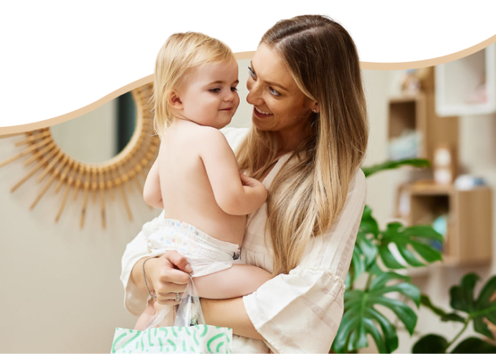 A smiling woman holding a baby in a diaper indoors.