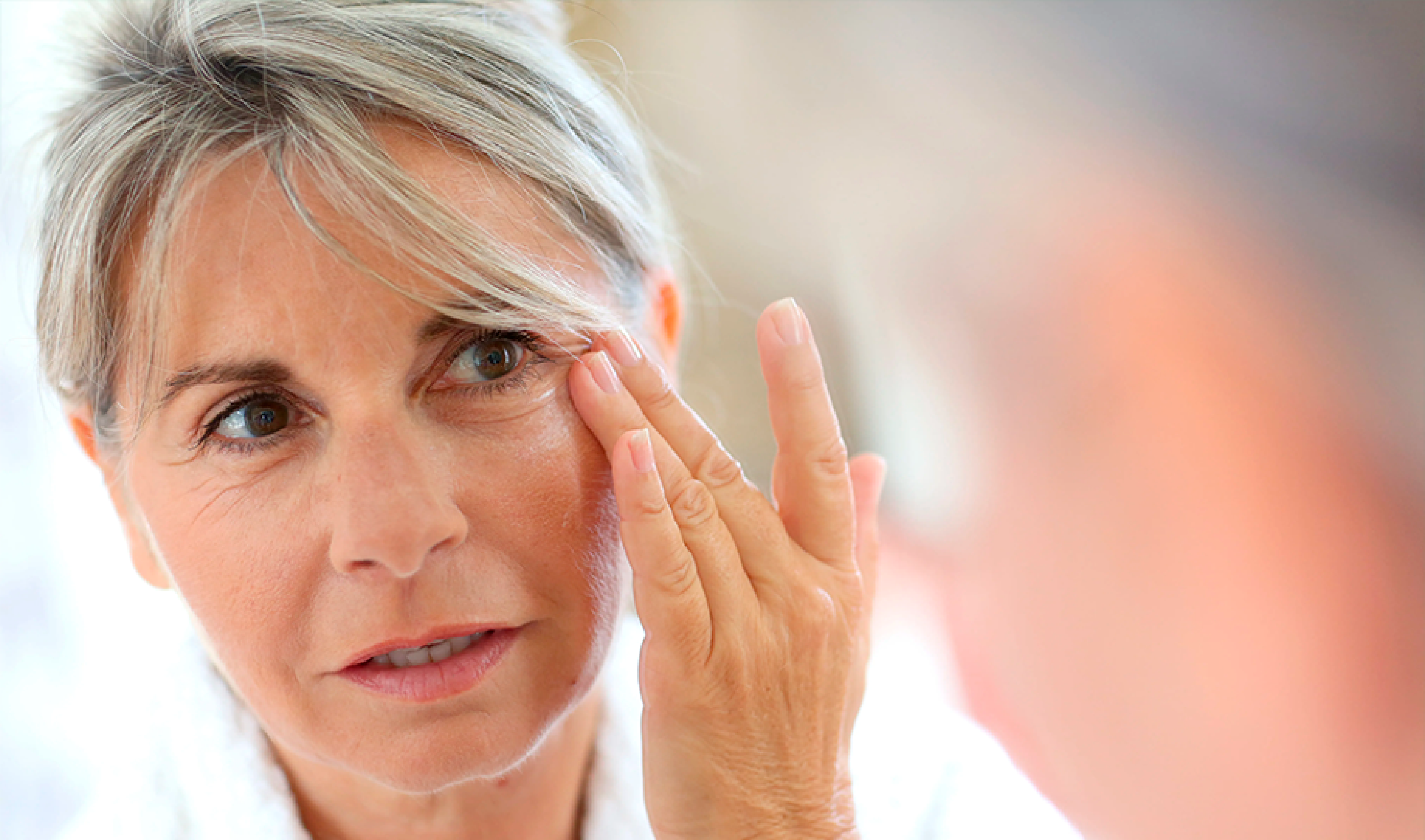 Woman applying skincare near her eye in front of a mirror.