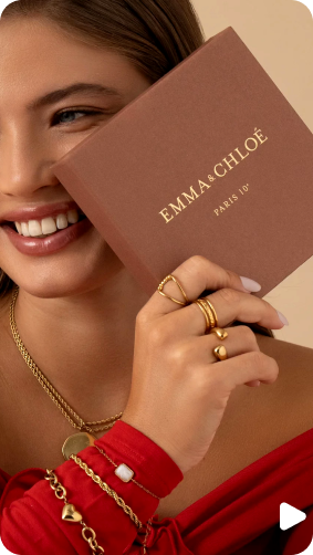 Woman holding an Emma & Chloé box, smiling, wearing gold jewelry and red top.