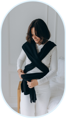 Woman adjusting a black baby wrap carrier across her shoulders in a bright room.