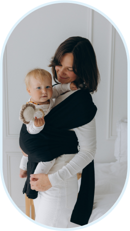 A woman holding a baby in a black baby carrier, standing in a white room.