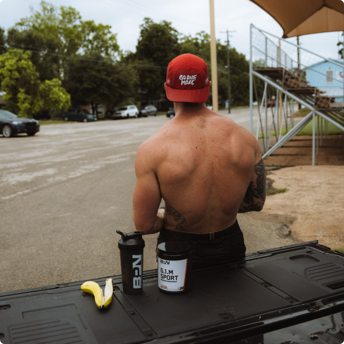 Shirtless man with a red cap sits outdoors beside workout supplements and a banana.