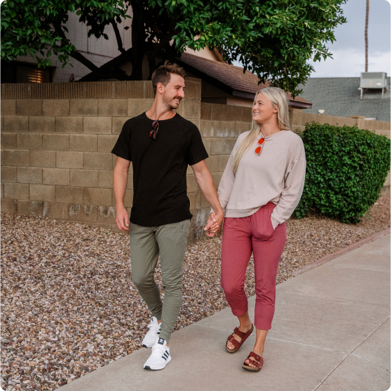 A couple holding hands while walking on a sidewalk, smiling at each other.