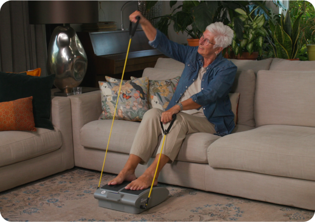 Elderly person exercising on a couch with a resistance band machine.