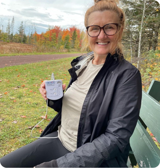 Woman sitting on a bench outdoors holding a medical device, with autumn foliage in the background.