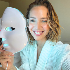 Smiling woman holding a LED therapy face mask in a robe.