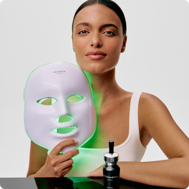 Person holding an LED facial mask and serum bottle, wearing a white top.