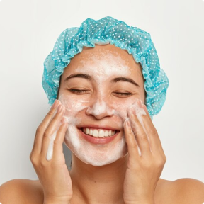 Person smiling while washing face, wearing blue shower cap.