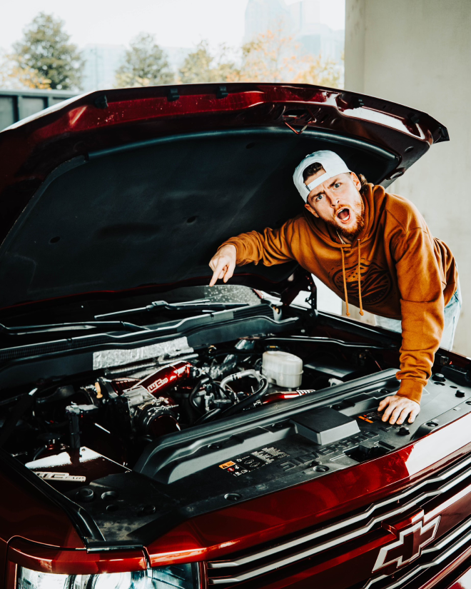 Person looking surprised under car hood, wearing orange hoodie and cap.