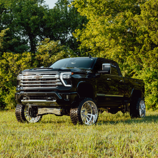 Lifted black truck with large chrome wheels parked on grass, trees in the background.