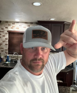 Man in a kitchen wearing a cap and white shirt, gesturing with his hand.
