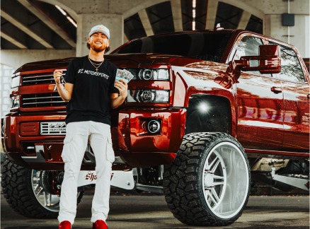 Man smiling beside a large, lifted, red truck under a bridge.