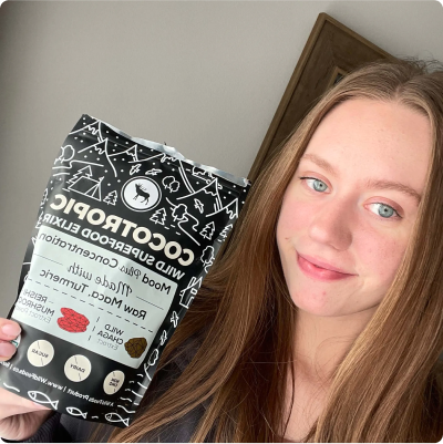 A person holding a package of 'Cocotropic, Wild Superfood Elixir,' smiling at the camera.