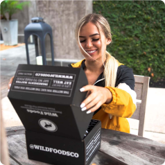 Person smiling while opening black box labeled 'WILD FOOD CO' outdoors at a table.