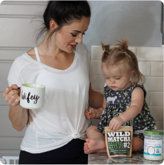 A woman holding a mug stands beside a child seated on a counter with matcha powder.